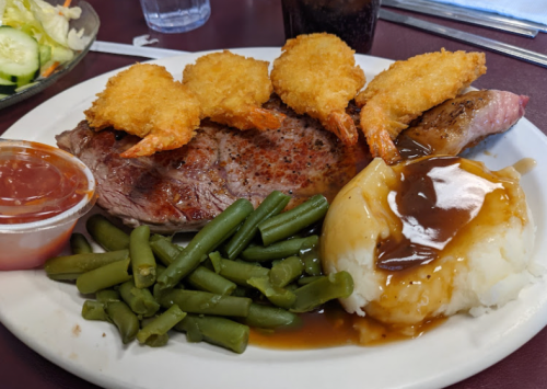 A plate with a steak, fried shrimp, green beans, and mashed potatoes with gravy, accompanied by a small cup of sauce.