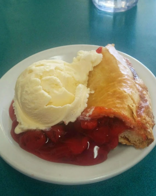 A slice of cherry pie topped with a scoop of vanilla ice cream on a white plate.