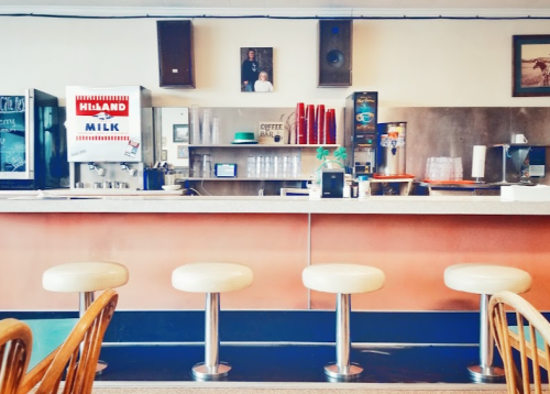 A retro diner interior featuring a counter with four stools, a milk dispenser, and vintage decor on the walls.