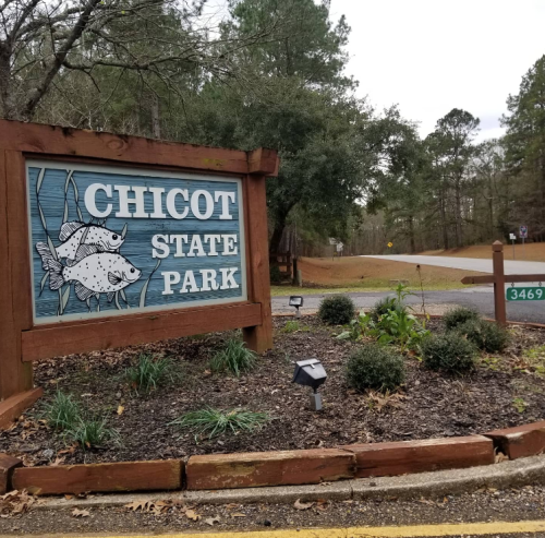 Sign for Chicot State Park featuring fish illustrations, surrounded by greenery and a gravel road in the background.