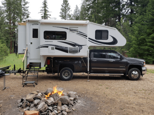 A truck with a camper mounted on top parked near a campfire in a forested area.