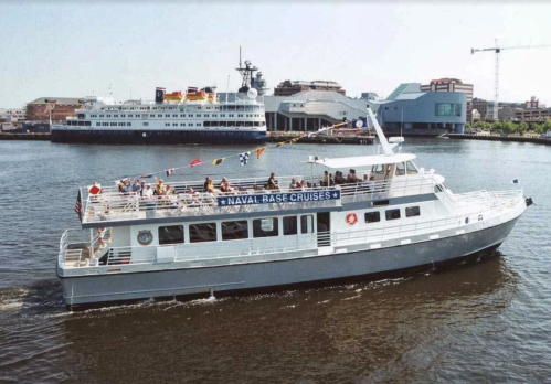 A white cruise boat with passengers sails on a calm waterway, with a larger ship and buildings in the background.