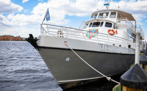 A close-up of the Victory Rover boat docked by the water, with a cloudy sky in the background.