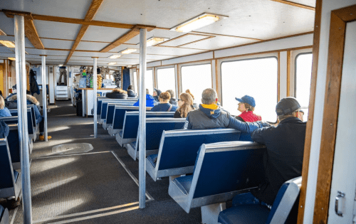 Interior of a ferry with passengers seated on blue benches, looking out large windows. Bright and spacious atmosphere.