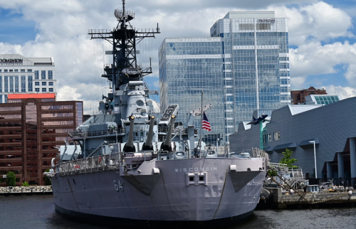 A battleship named Wisconsin docked near modern buildings and a waterfront in a city setting.