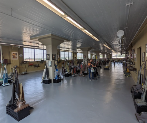 A spacious room filled with various vintage vacuum cleaners displayed along the walls, with a person examining them.