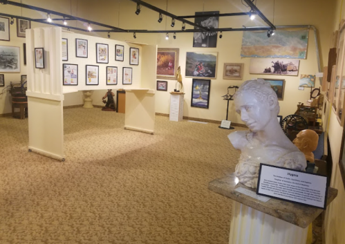 A spacious art gallery featuring framed artworks, sculptures, and a bust on a pedestal with an informational plaque.