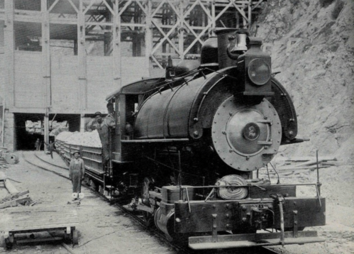 A vintage steam locomotive at a construction site, with a worker standing beside it and wooden structures in the background.