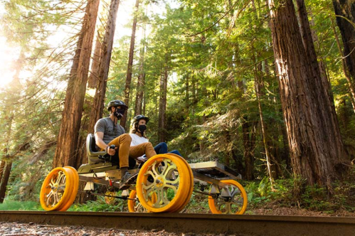 Two people ride a unique four-wheeled vehicle on a forest path surrounded by tall trees.