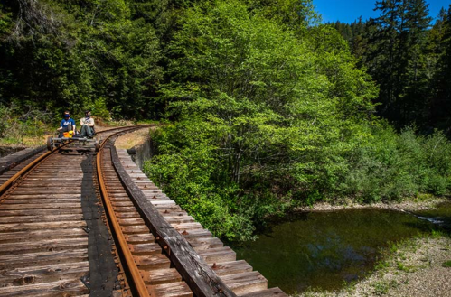 Two people on a small rail cart on wooden tracks beside a lush green forest and a calm pond.