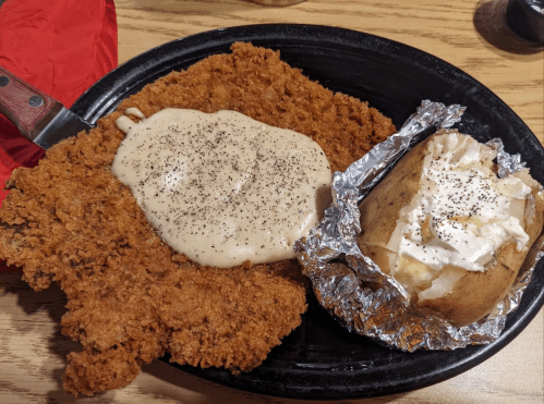 A plate with a large piece of fried chicken topped with gravy, alongside a baked potato with sour cream and chives.