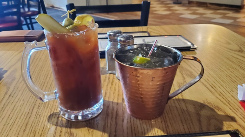 Two cocktails on a wooden table: a Bloody Mary in a glass and a mixed drink in a copper mug.
