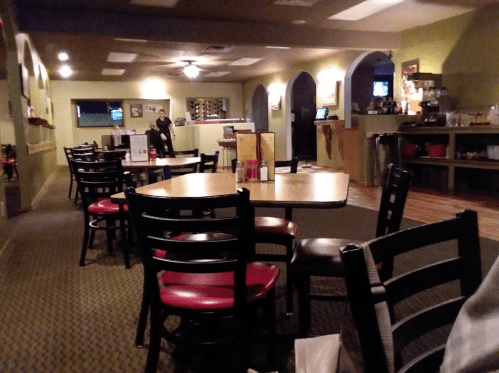 Interior of a restaurant with empty tables and chairs, a bar area in the background, and a staff member at the counter.