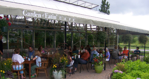 A café with outdoor seating surrounded by plants, where people are enjoying meals and drinks under a canopy.