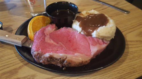 A plate with a large slice of pink prime rib, mashed potatoes with gravy, an orange slice, and a small cup of sauce.