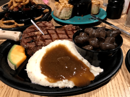 A plate featuring a grilled steak, mashed potatoes with gravy, sautéed mushrooms, and a slice of cantaloupe.