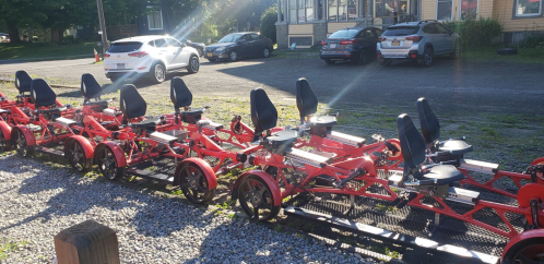 A row of red pedal-powered vehicles with black seats parked on a gravel lot, with cars and a building in the background.