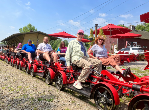 A group of people sitting on red pedal-powered rail cars under sunny skies, with umbrellas providing shade.
