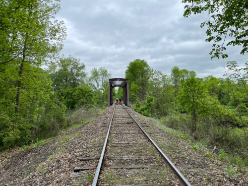 A view of a railway track leading to a bridge, surrounded by lush green trees under a cloudy sky.