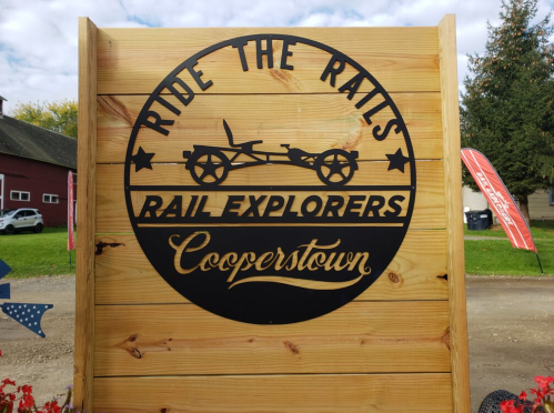 Sign for "Ride the Rails Rail Explorers" in Cooperstown, featuring a vintage rail cart design on a wooden background.