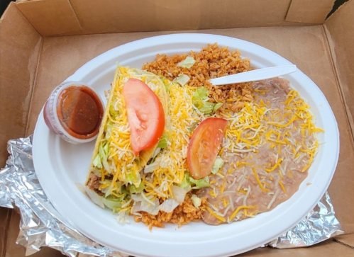 A takeout plate with two tacos topped with lettuce, cheese, and tomato, served with rice and refried beans.