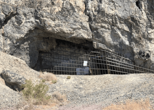 A rocky cliff with a cave entrance, partially reinforced with metal rebar and surrounded by dry vegetation.