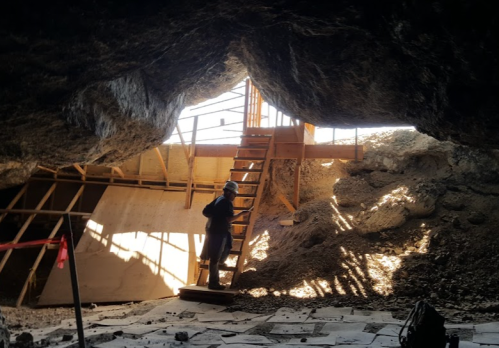 A person stands inside a cave, next to a wooden staircase leading up, with rocky walls and construction materials around.