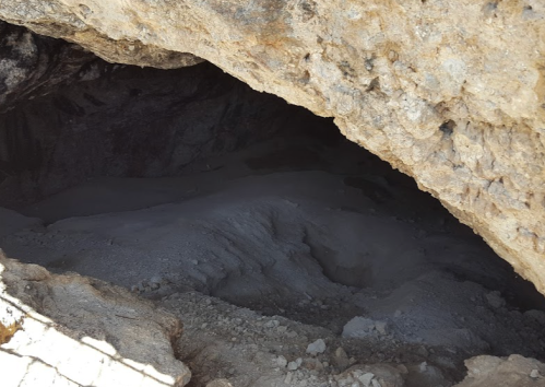 A dark cave entrance with rocky walls and a sandy floor, partially illuminated by natural light.