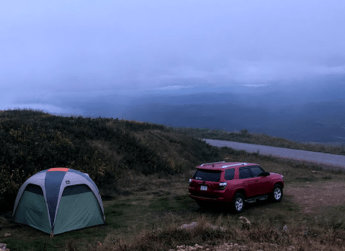 A red SUV parked beside a tent on a misty hillside, with a cloudy sky and distant mountains in the background.
