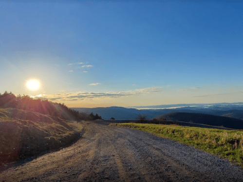 A gravel road winds through a scenic landscape at sunset, with rolling hills and a clear blue sky.