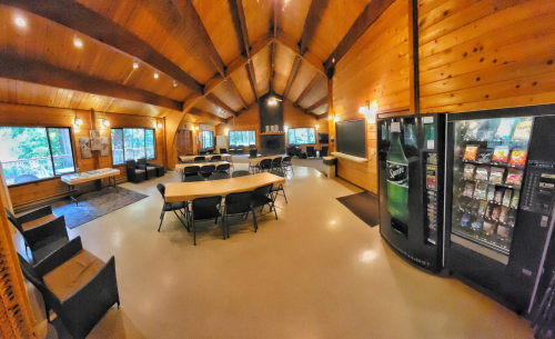 A spacious wooden interior with tables, chairs, a vending machine, and large windows overlooking greenery.