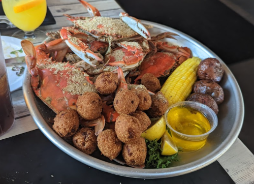 A metal platter filled with crabs, corn on the cob, hushpuppies, and a lemon wedge, accompanied by a small cup of sauce.