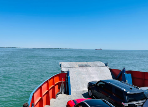 A view from a ferry showing a ramp leading to the water, with cars parked on the deck and a clear blue sky.