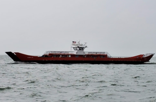 A red ferry boat sails on calm waters under a gray sky, with an American flag flying from its deck.