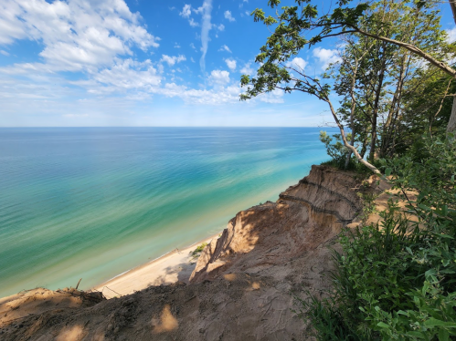 A scenic view of a sandy beach and calm blue waters, framed by trees and a clear sky with scattered clouds.