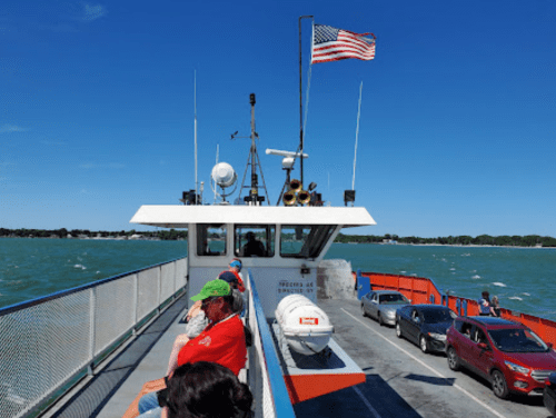 View from a ferry deck with passengers, an American flag, and vehicles on a sunny day with blue water.