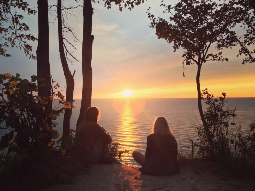 Two people sit on a sandy shore, watching a vibrant sunset over a calm lake, framed by trees.