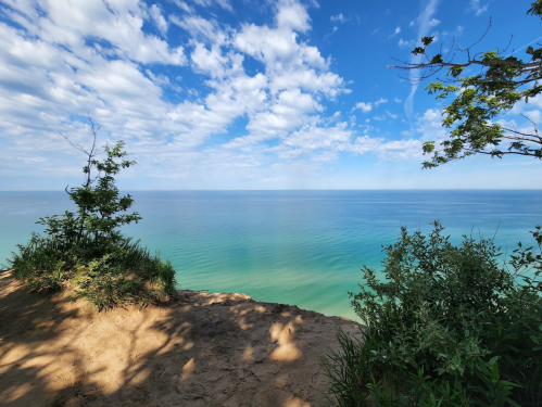 A serene view of a calm lake under a blue sky with scattered clouds, framed by greenery on the shore.