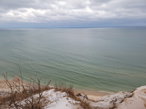 A serene view of a calm lake with gentle waves, surrounded by a sandy shore and cloudy skies.