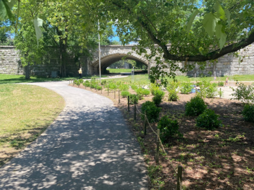 A sunny park scene featuring a winding path, greenery, and a stone arch bridge in the background.