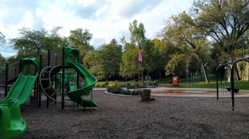 A playground with green slides and swings, surrounded by trees and a splash pad in a park setting.