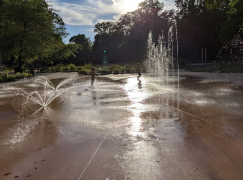 Children play in a splash pad with water fountains, surrounded by greenery and sunlight.