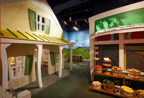 Colorful play area featuring a whimsical house and a market stand with fruits and vegetables. Bright, cheerful decor.