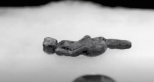 A small, abstract metallic figure lying on a white surface, partially obscured by a soft, blurred background.