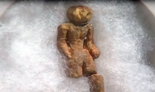 A small, ancient figurine made of stone, displayed on white cushioning material.