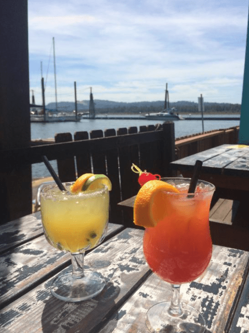 Two colorful cocktails on a wooden table by the water, with boats and mountains in the background under a clear sky.