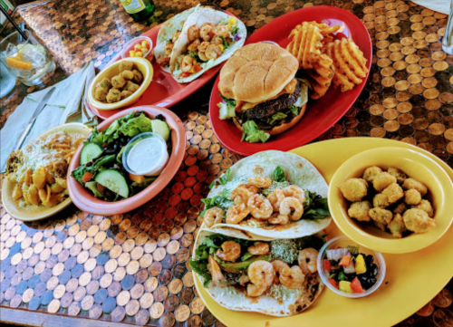 A colorful spread of seafood tacos, a burger with fries, and salads on a table with a shiny penny surface.