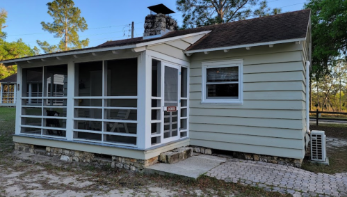 A small, light-colored house with a screened porch and stone foundation, surrounded by trees and grass.
