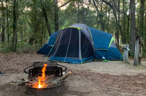 A blue and green tent set up in a wooded area, with a campfire burning in a metal fire pit nearby.