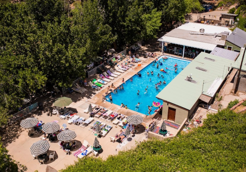 Aerial view of a vibrant pool area with people swimming, sunbathing, and lounging under umbrellas.